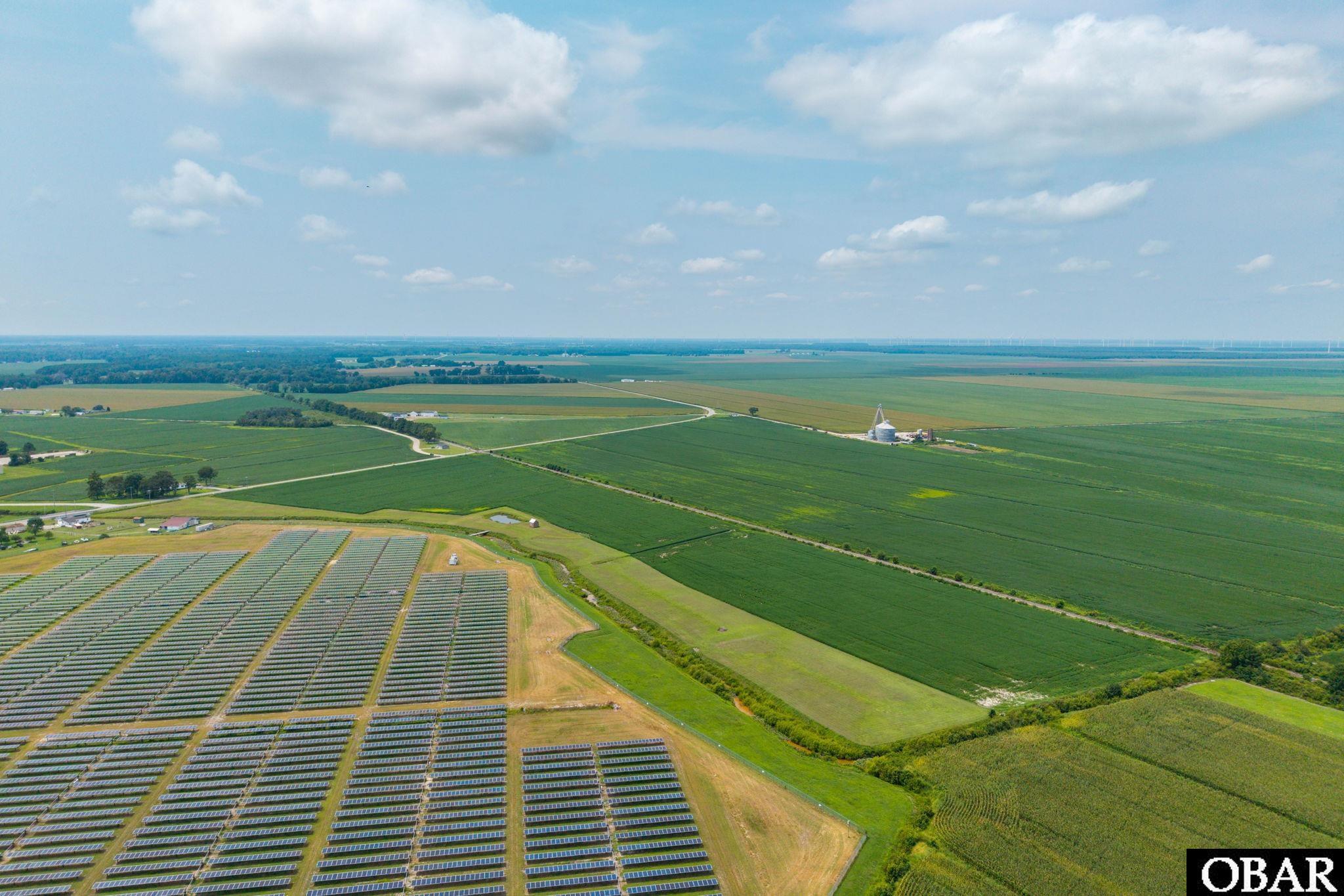 Aerial view of a solar array beside expansive green farmland and distant wind turbines.