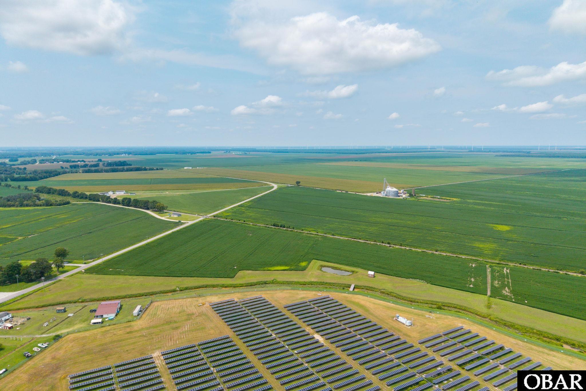Aerial view of a solar panel installation surrounded by green fields and distant wind turbines.