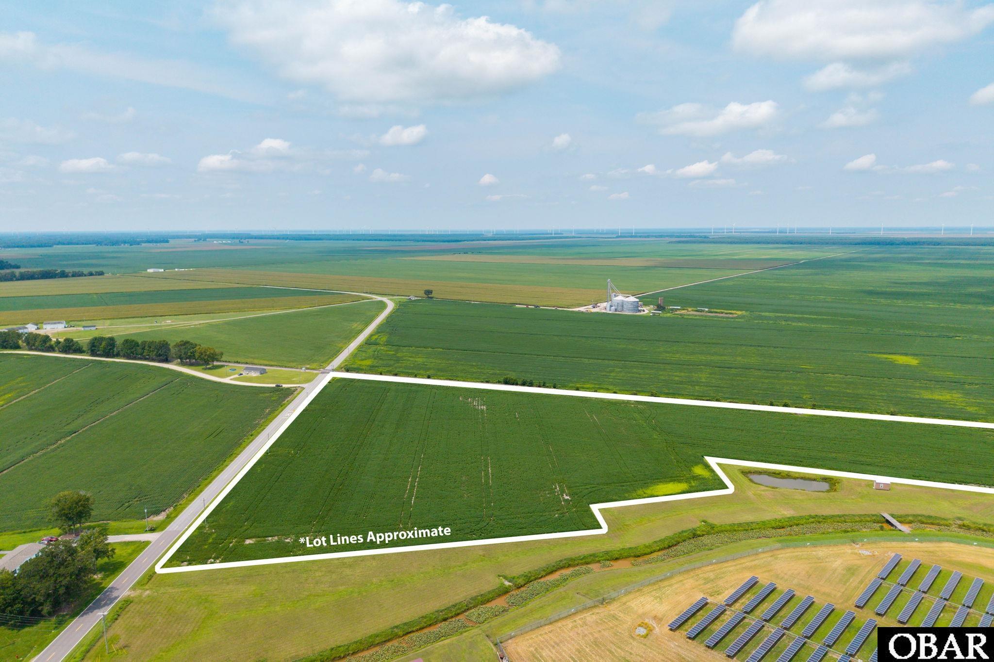 Aerial view of outlined farmland beside a road, with solar panels and distant wind turbines.