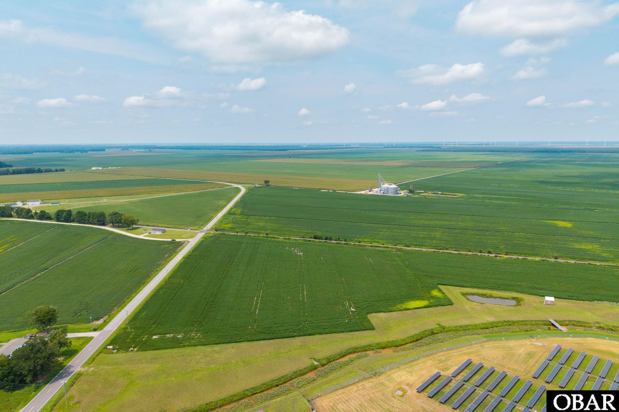 Aerial farmland with intersecting roads, a pond, foreground solar panels, and distant wind turbines.