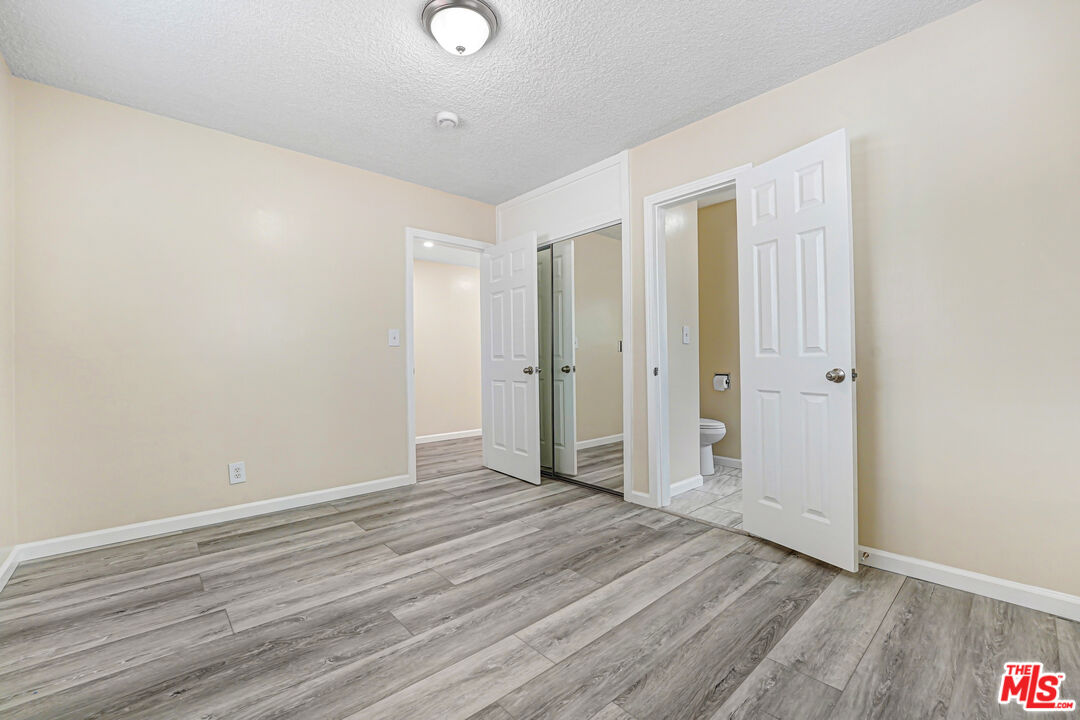 Empty bedroom with gray wood-look flooring, mirrored closet doors, and an adjoining bathroom.