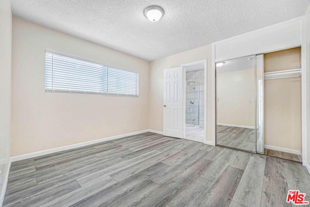 Empty bedroom with gray plank flooring, wide window with blinds, and mirrored closet doors.