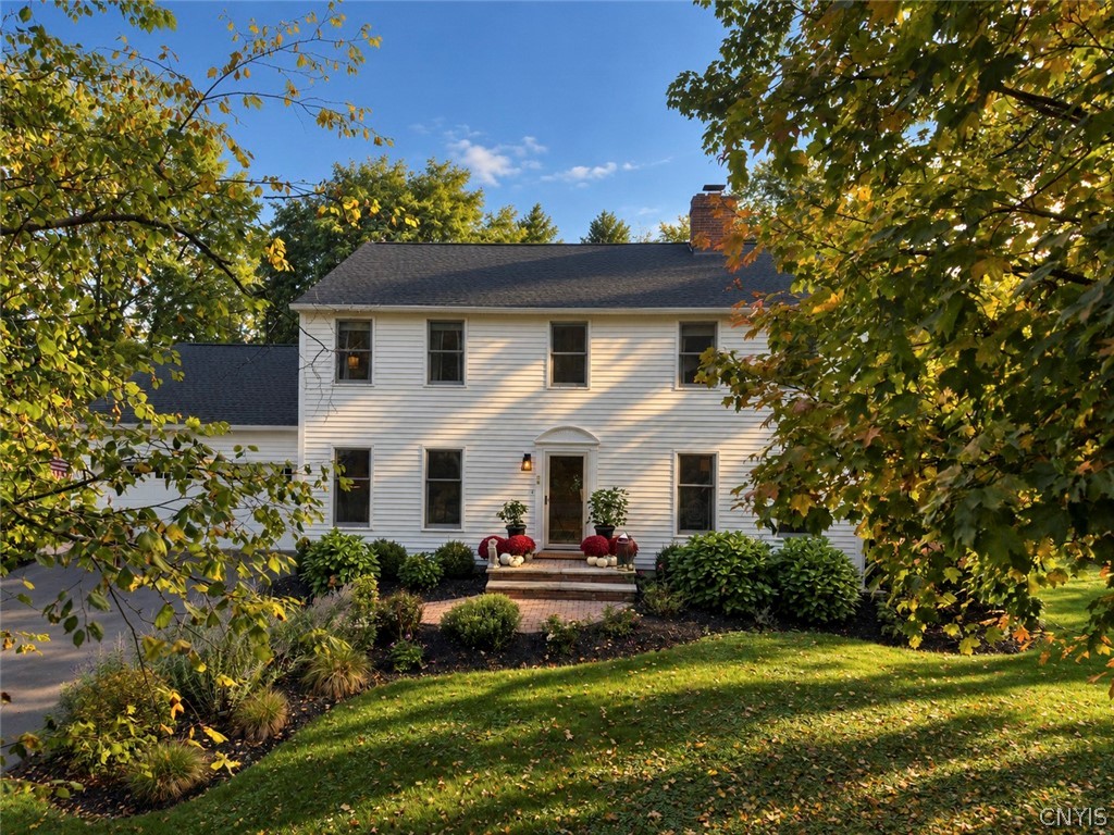 White two-story colonial home framed by autumn trees, with an attached garage and landscaped front garden.