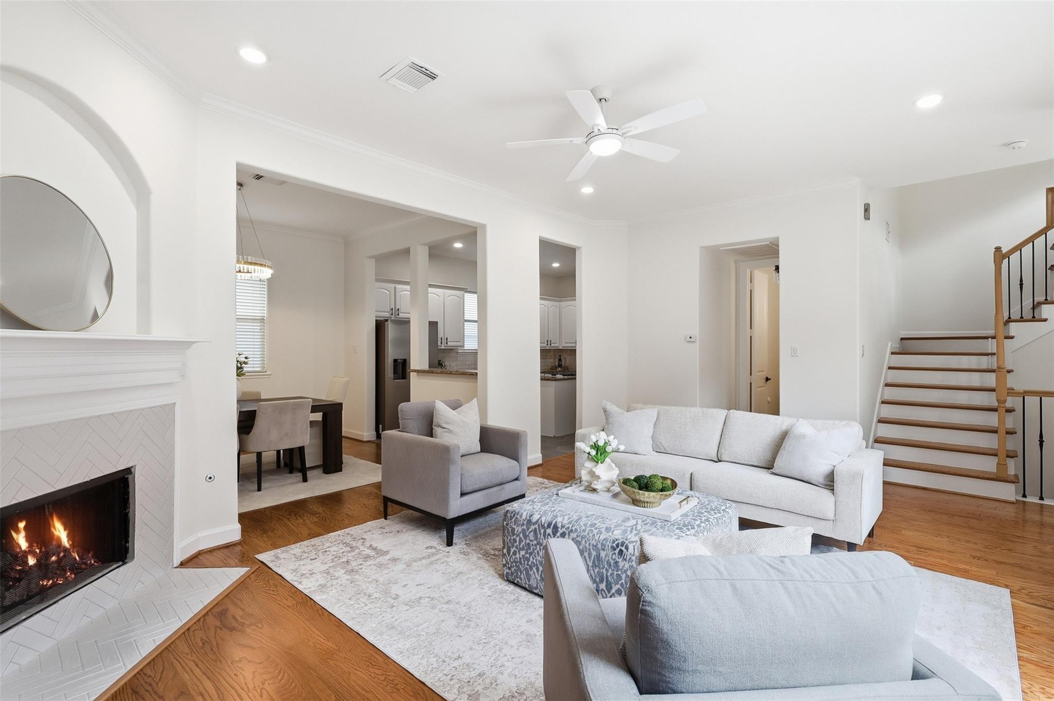 Staged living room with a lit tiled fireplace, hardwood floors, and an open view to the kitchen and staircase.