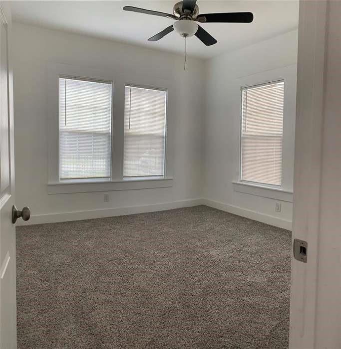 Empty bedroom with gray carpet, three windows with blinds, and a ceiling fan.