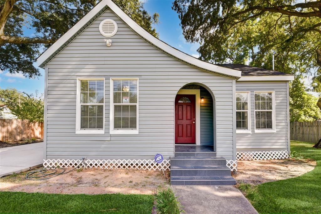 Gray single-story house with a red front door beneath an arched entry and mature trees overhead.