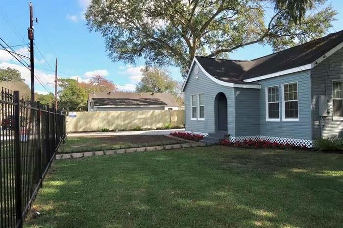 Gray-blue house with an arched entry, flower bed, fenced lawn, and paved drive.