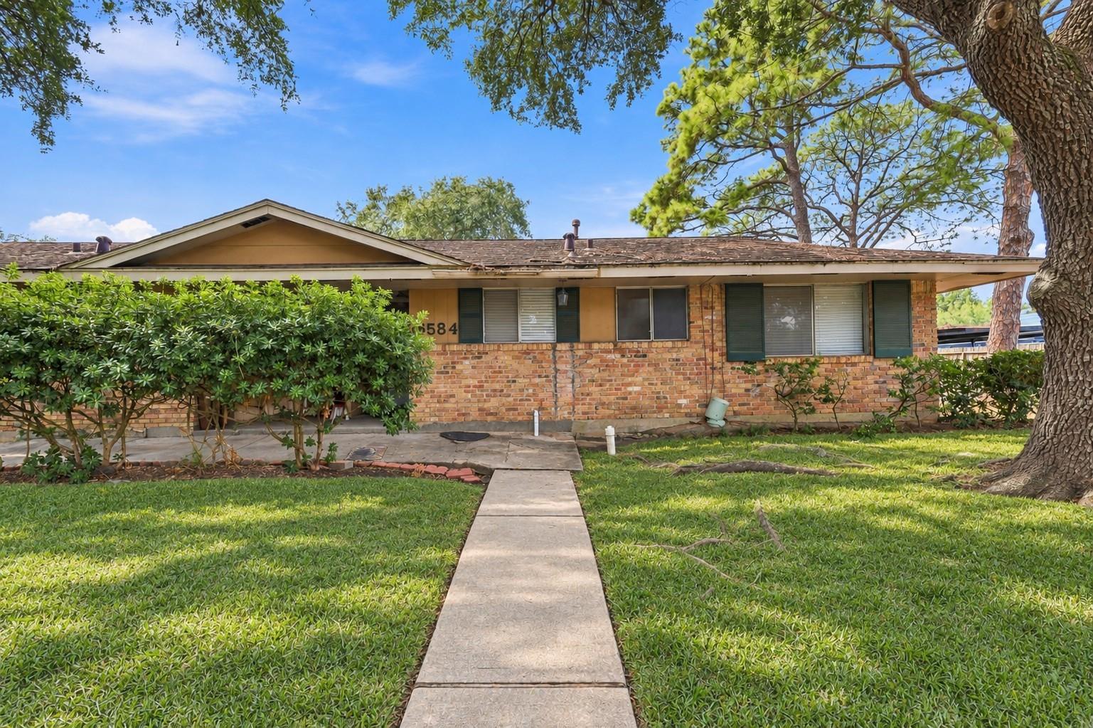 Single-story brick house with mature trees and a concrete walkway across the lawn.