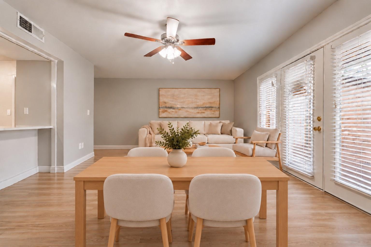 Staged dining and living area with a wooden table, neutral seating, and glass doors with white blinds.