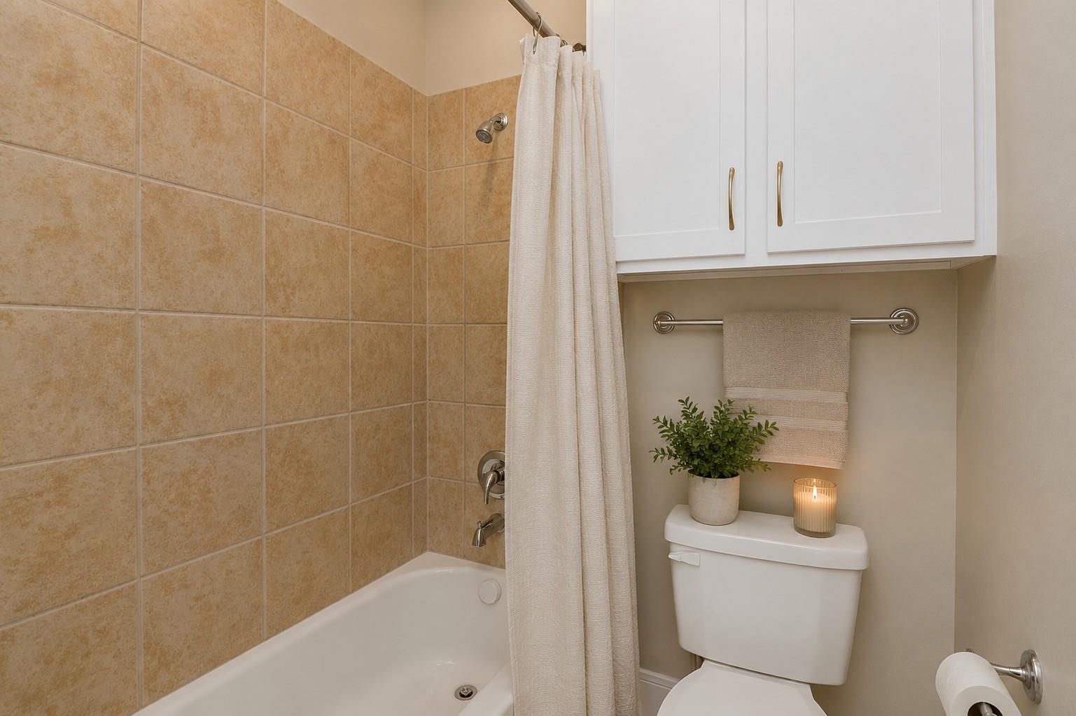 Bathroom with tan tiled tub and shower, white cabinets, and a decorated toilet tank.