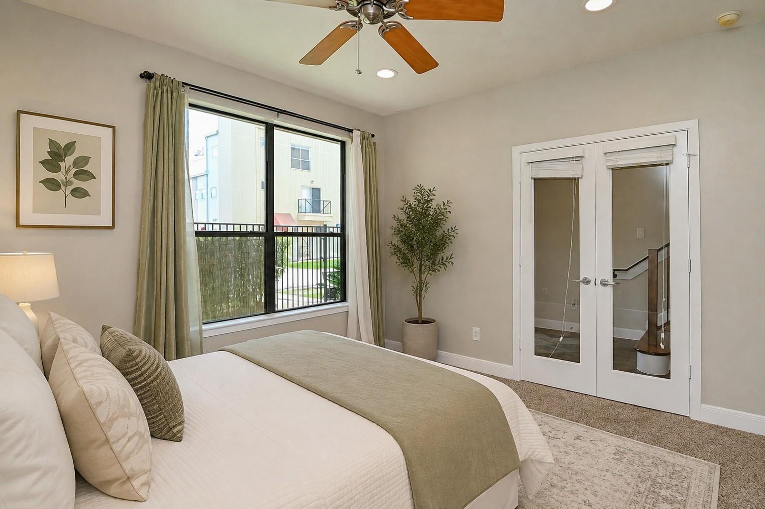 Staged bedroom with neutral bedding, a ceiling fan, and a large window overlooking a balcony.