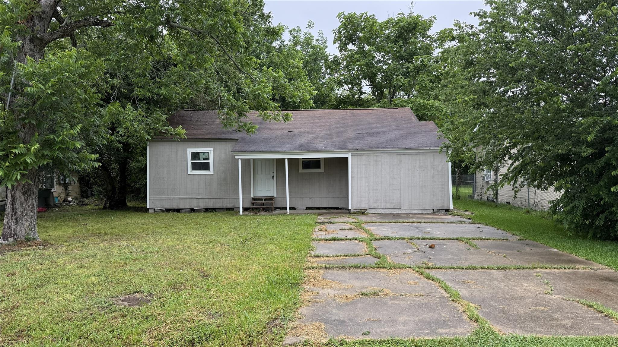 Gray single-story house with a covered porch, mature trees, and a cracked concrete driveway.
