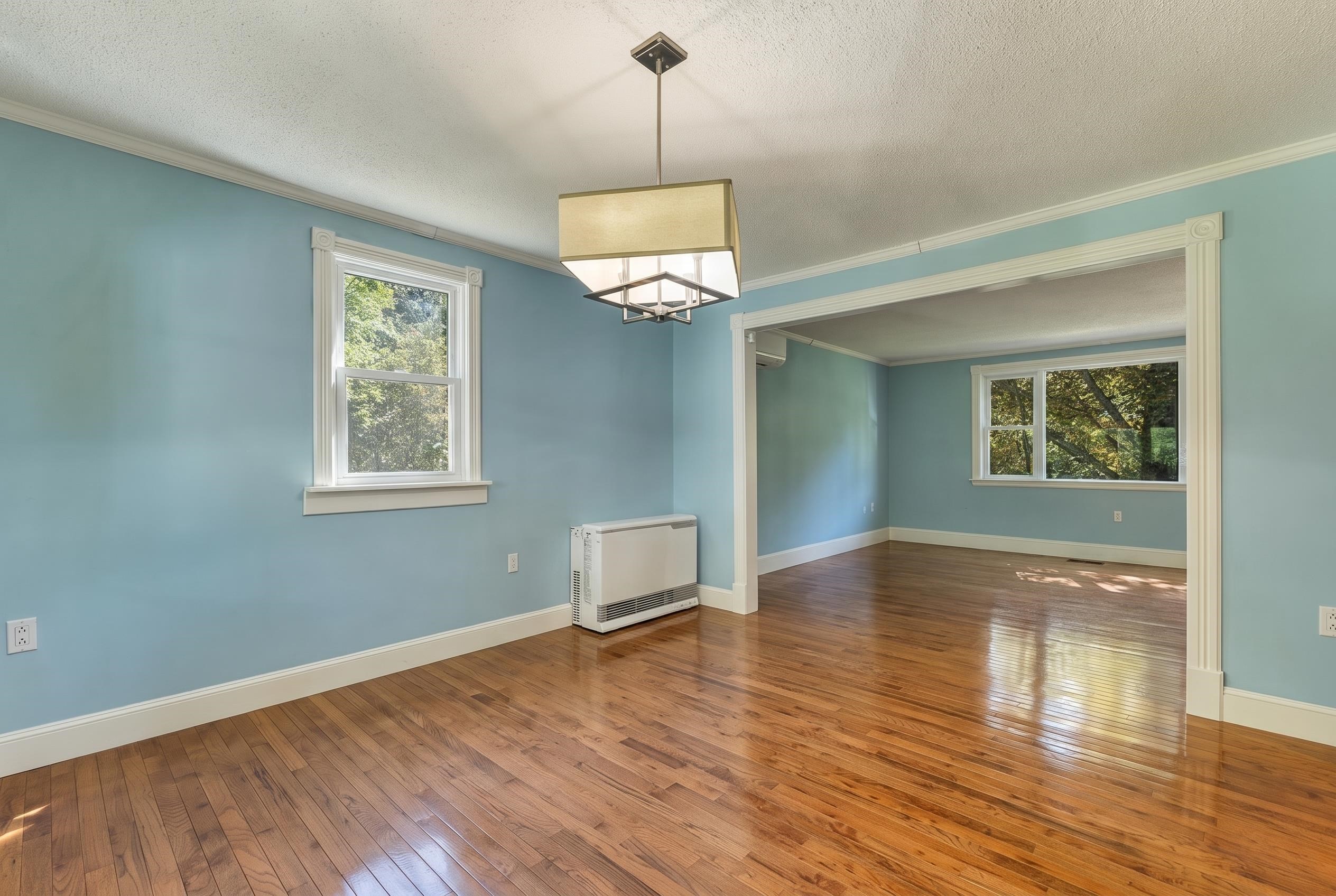 Unfurnished blue room with polished hardwood floors and a wide opening to a sunlit adjoining room.