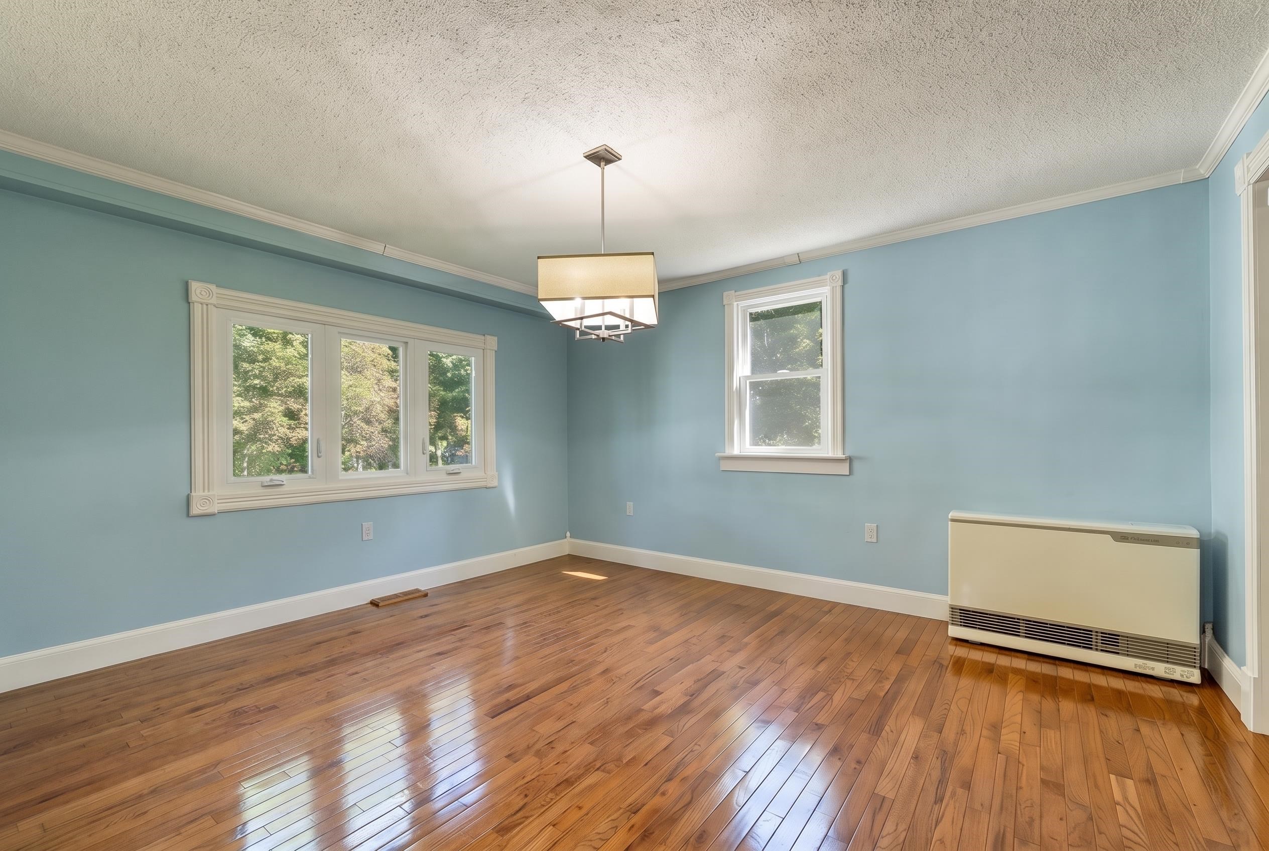 Empty blue-walled room with polished hardwood floors, large windows, and a wall-mounted heater.