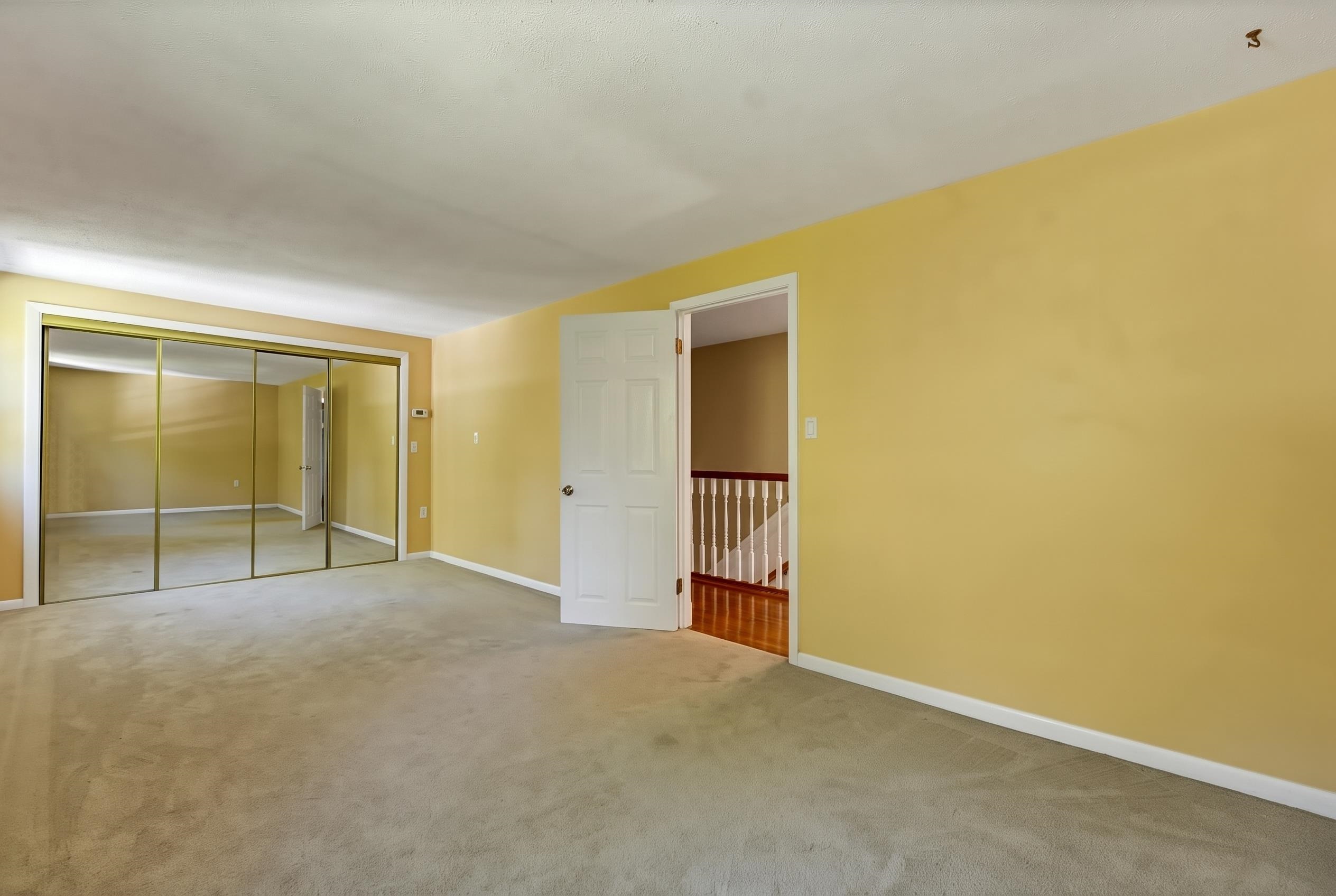 Empty yellow bedroom with beige carpet, mirrored closet doors, and an open door to the staircase.