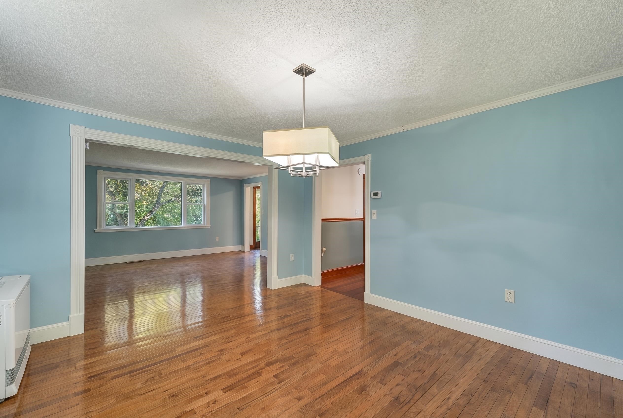Empty blue-walled dining area with polished hardwood floors, a square pendant light, and an adjoining windowed room.