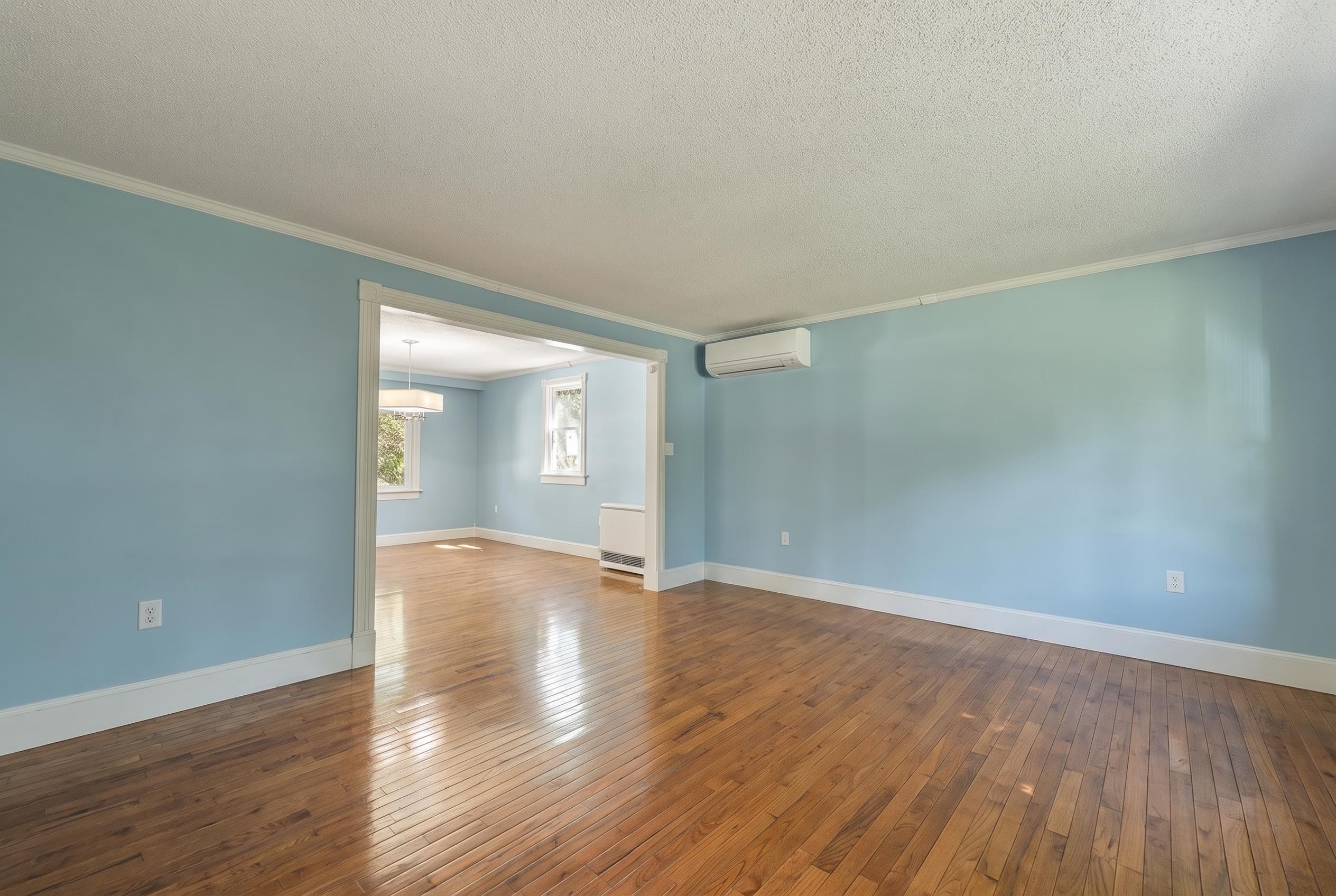 Empty blue room with hardwood floors, a wall-mounted mini-split, and a wide opening to a sunlit room.