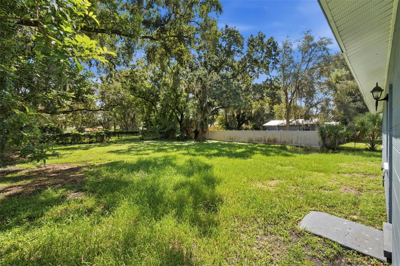 Grassy backyard with mature shade trees, a white fence, and a concrete walkway beside the house.