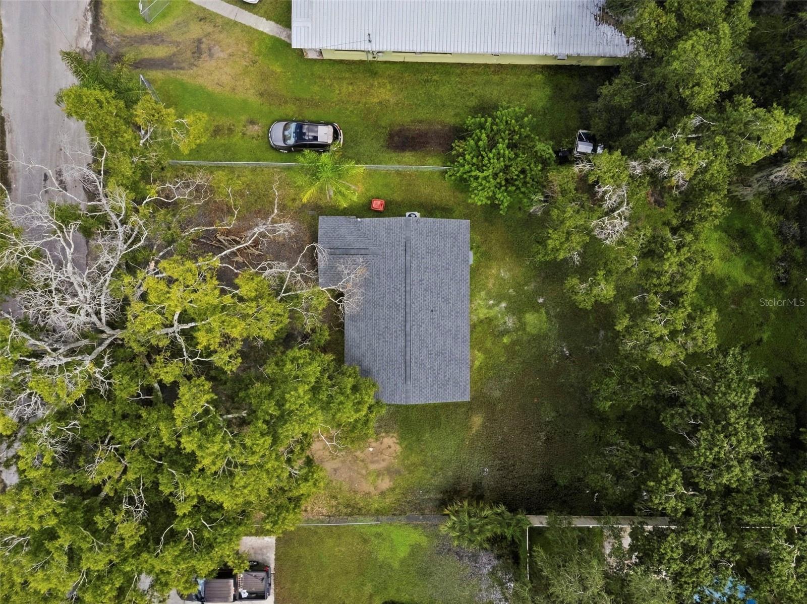 Aerial view of a gray-roofed house in a tree-filled yard beside a road.
