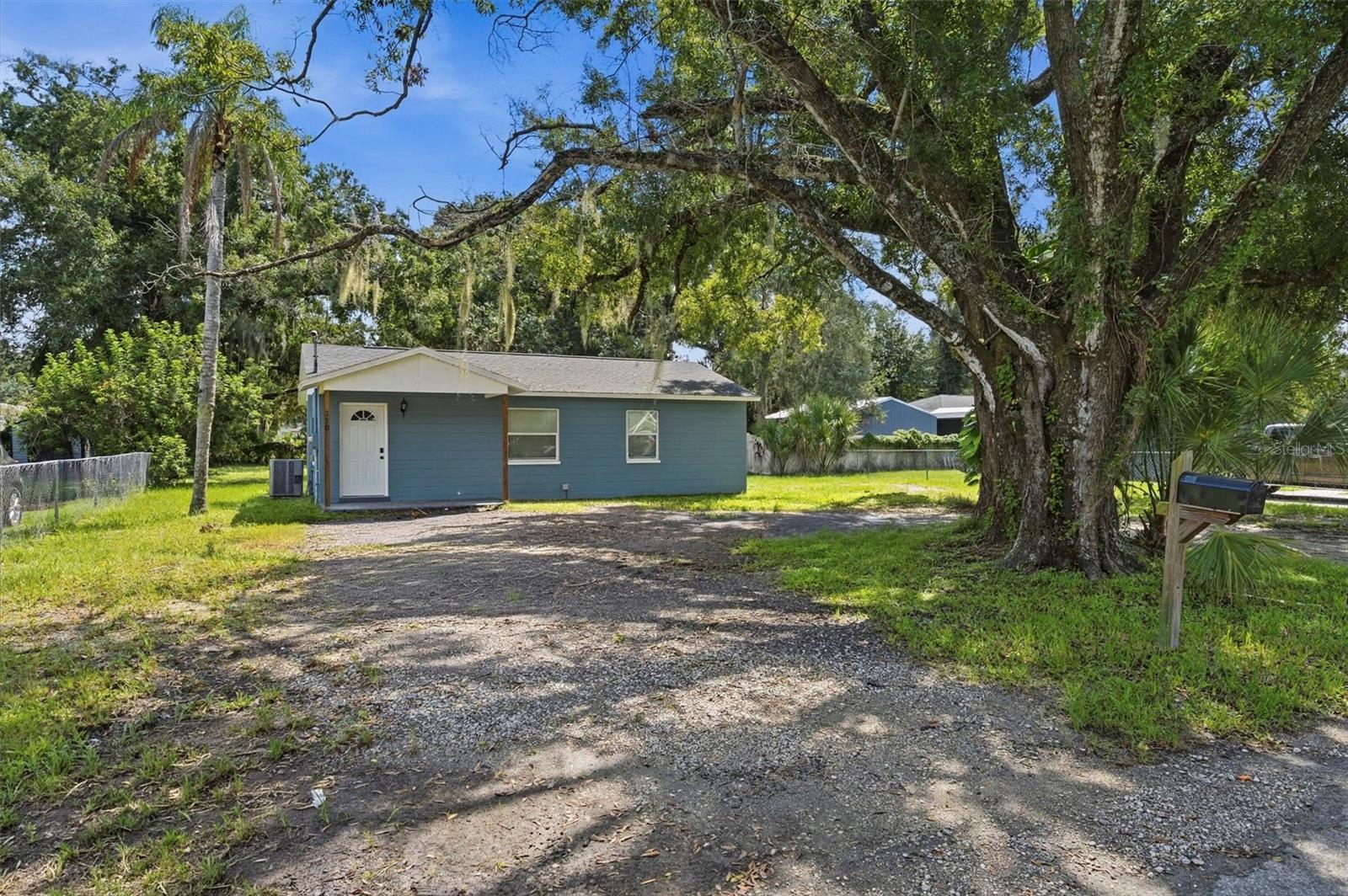 Blue-sided house with a gravel driveway beneath a large oak tree.