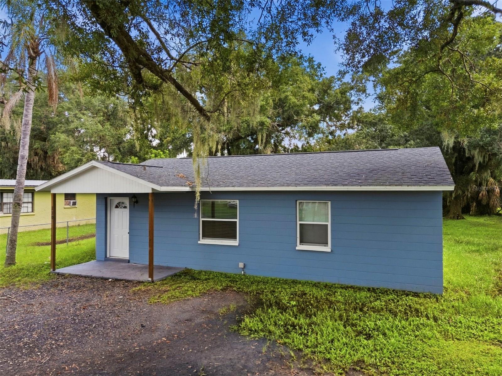 Blue single-story house with a covered entry, paved driveway, and mature trees.