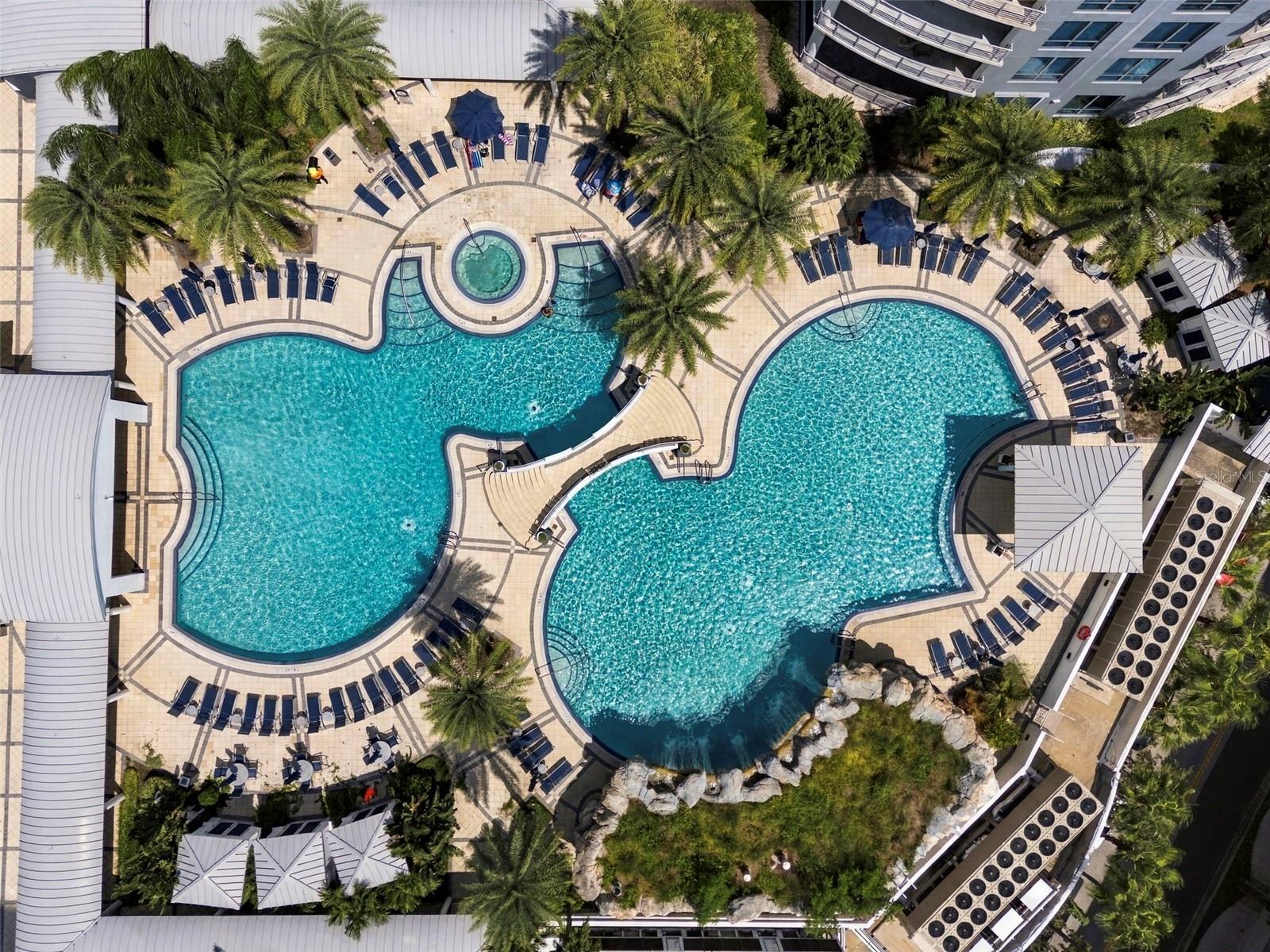 Aerial view of two curving condominium pools surrounded by lounge chairs and palm trees.
