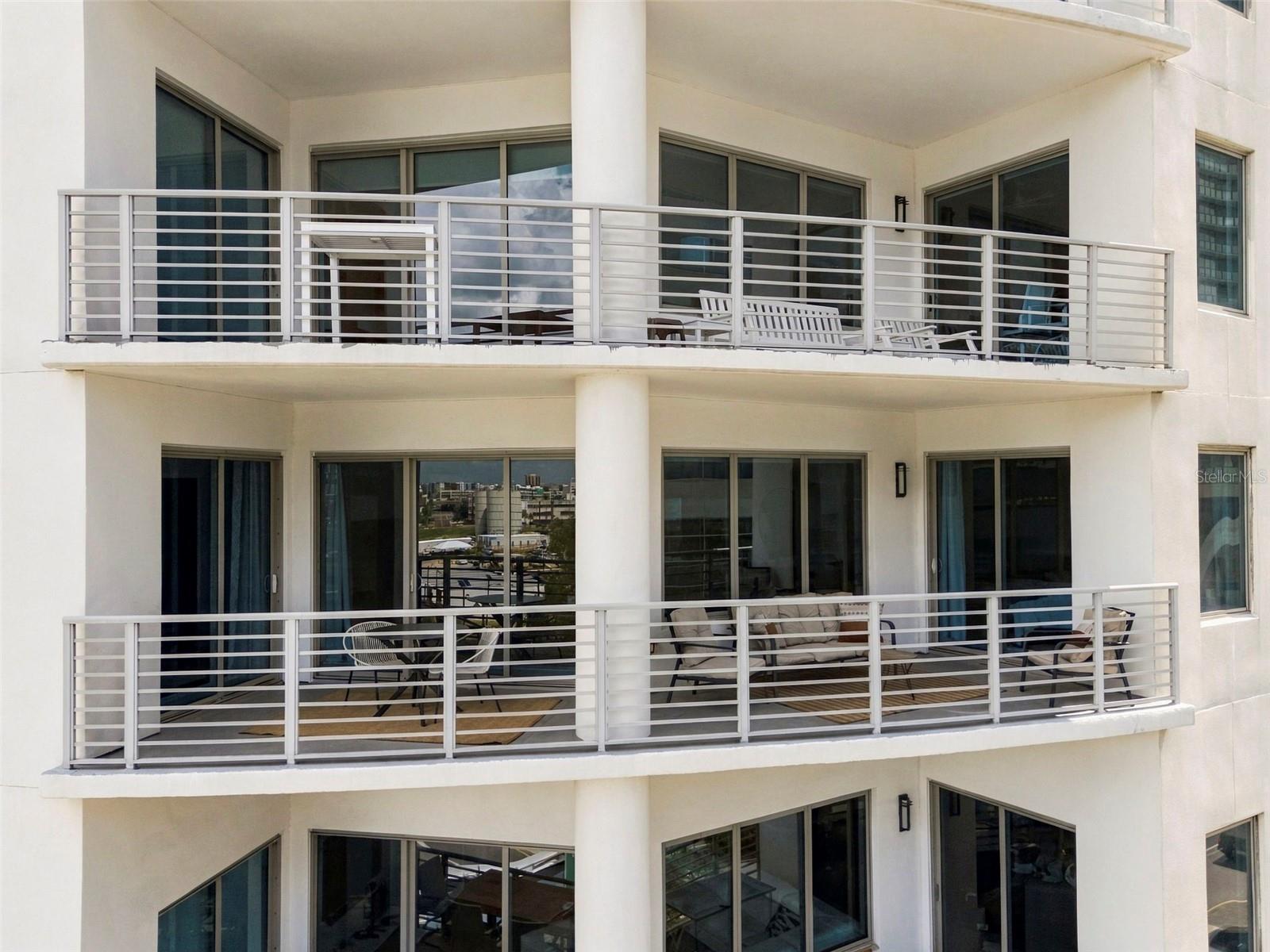 Modern white condominium facade with two furnished balconies and large sliding glass doors.
