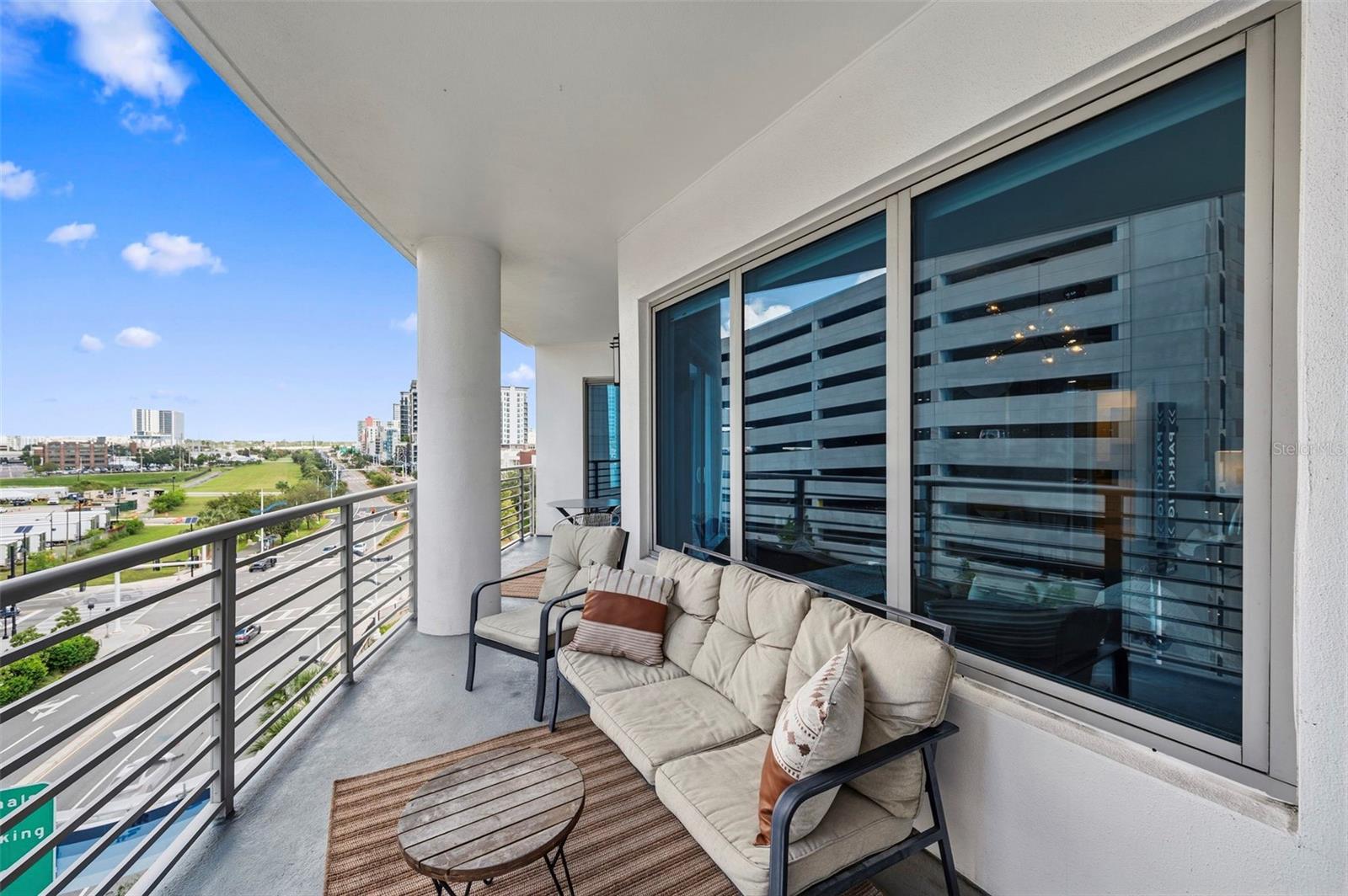 Condominium balcony furnished with cushioned seating and a coffee table, overlooking city streets and buildings.