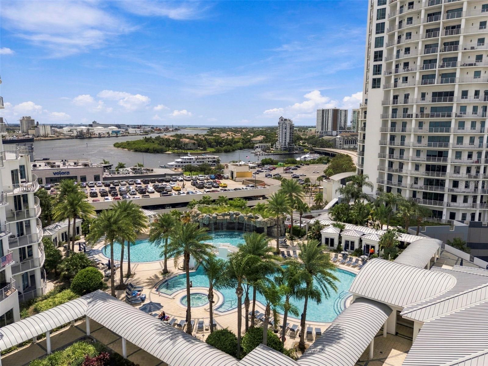 Condominium pool deck with palm trees and covered walkways overlooking a river and city buildings.