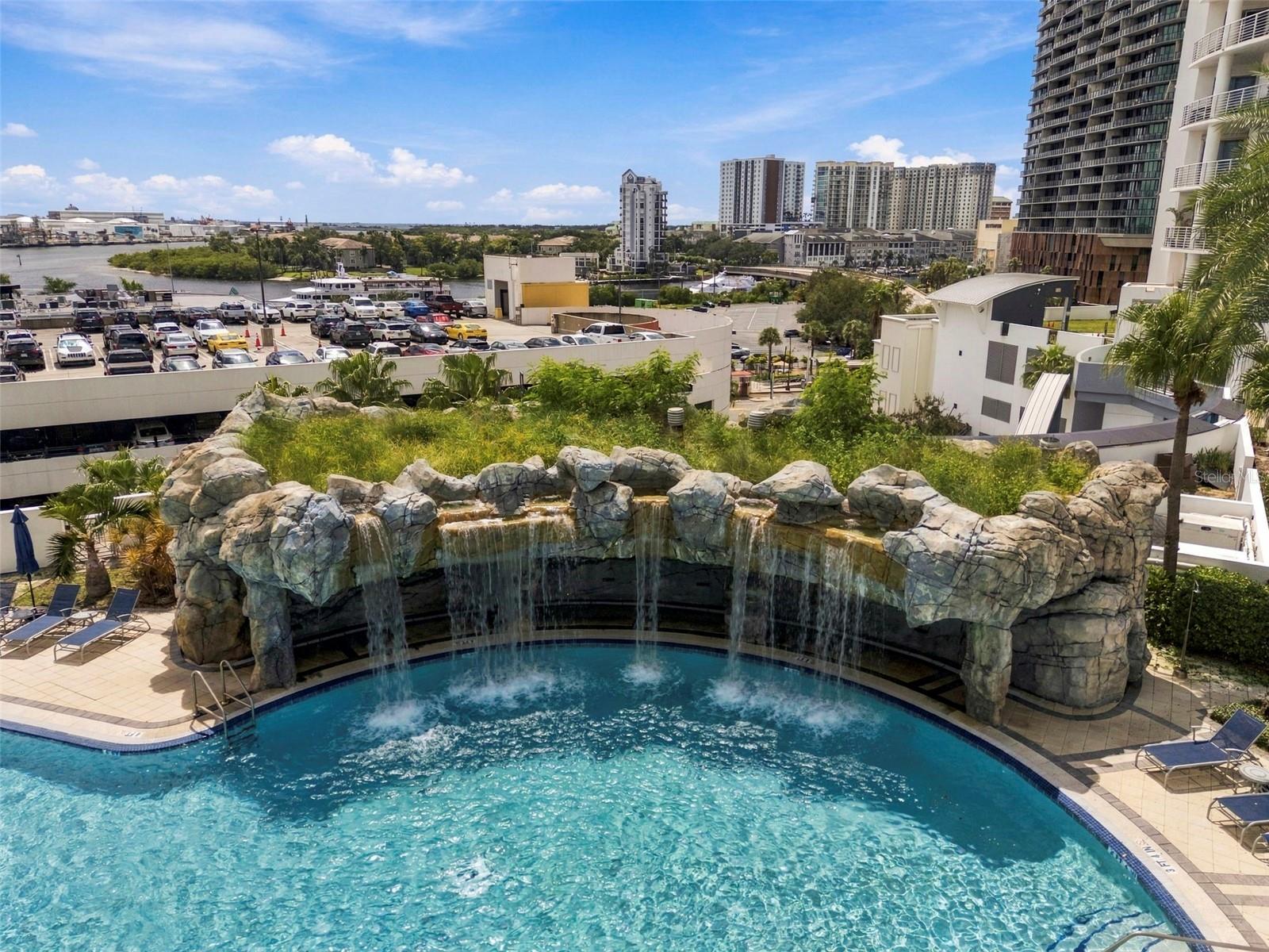 Elevated view of a pool with a rock waterfall and city buildings beyond.