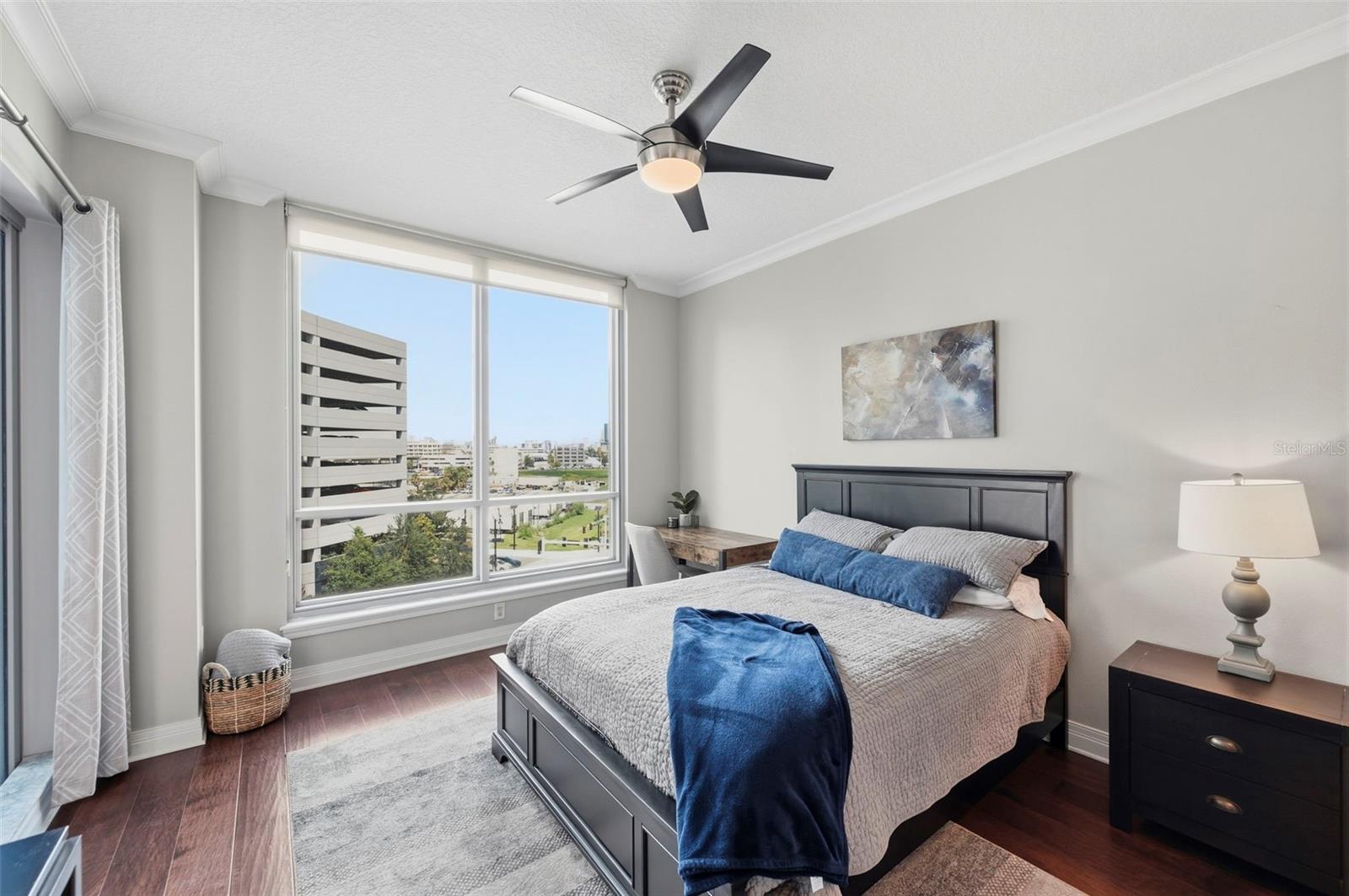 Bedroom with a dark wood bed, blue pillows, and large windows overlooking nearby buildings.