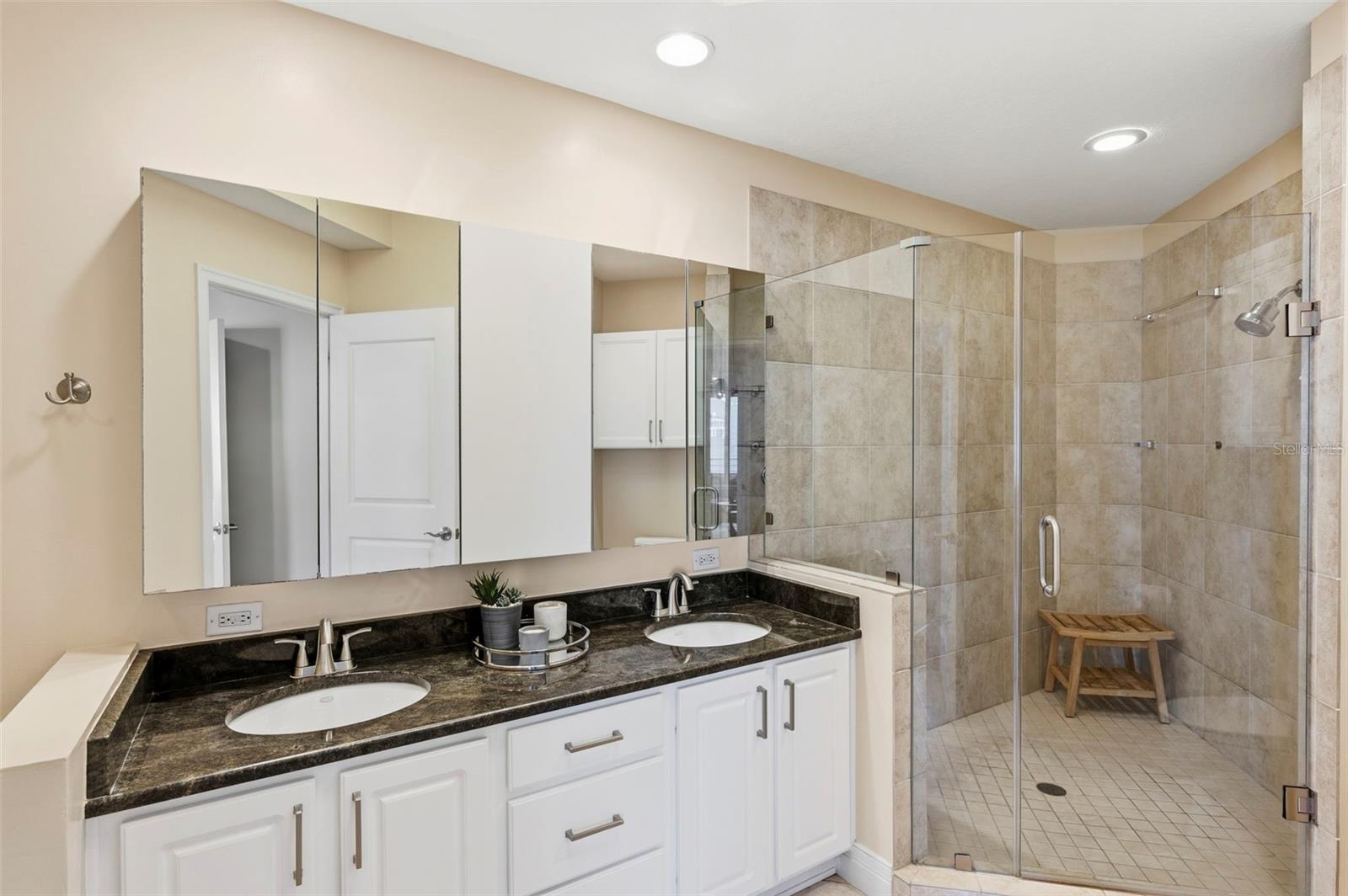 Bathroom with a double-sink vanity and glass-enclosed tiled shower with a wooden bench.