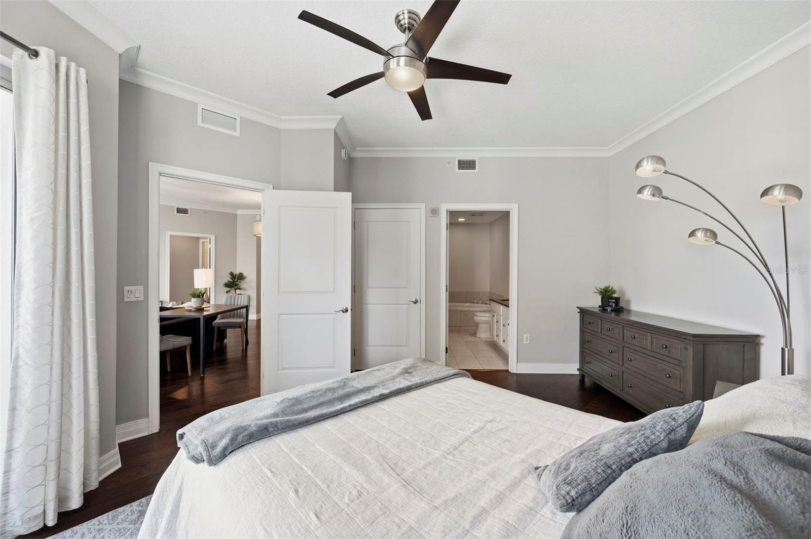 Furnished bedroom with a dark ceiling fan, dresser, and open doorways to the dining area and bathroom.