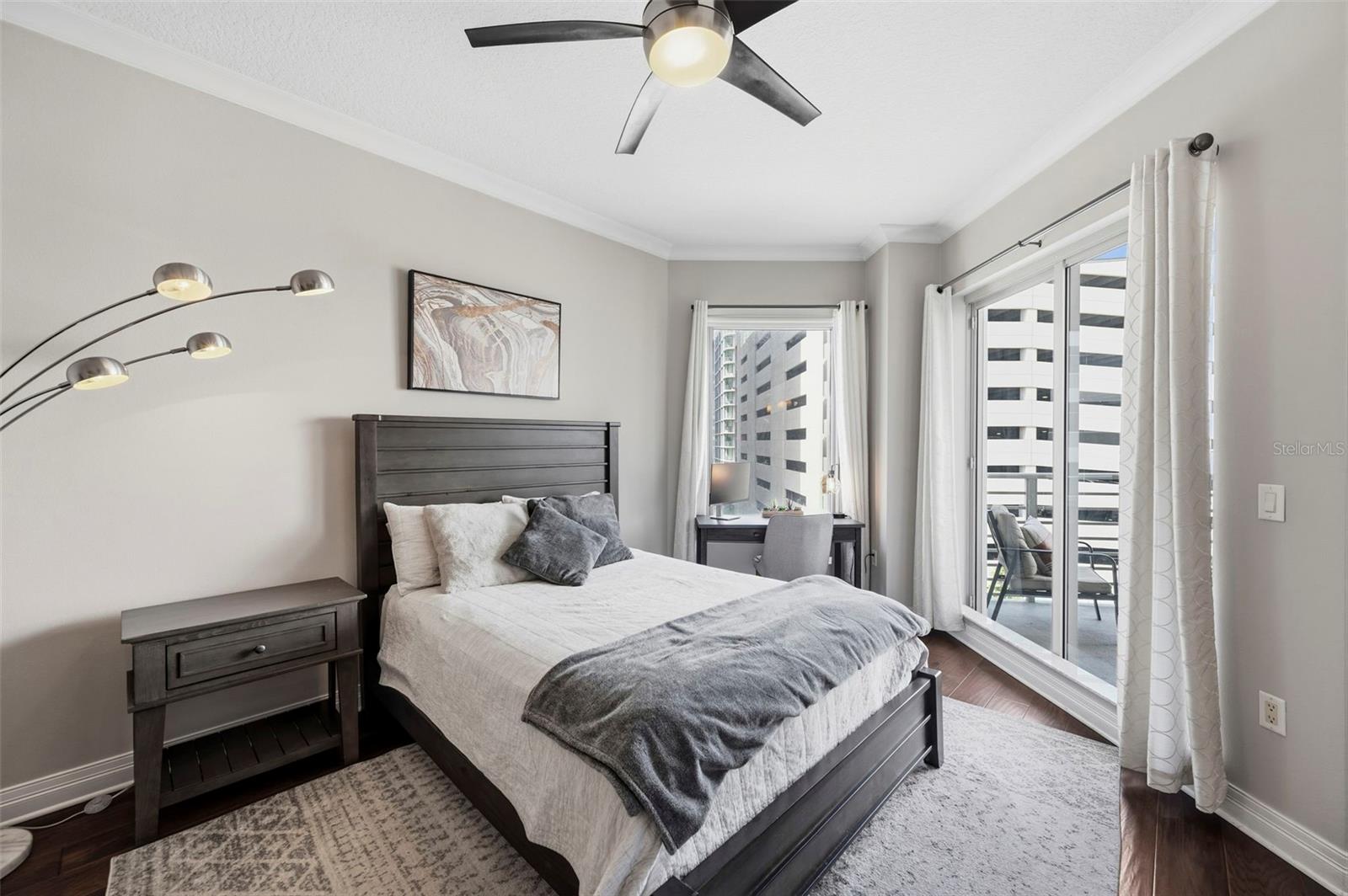Staged bedroom with a dark wood bed, desk, and balcony windows overlooking nearby buildings.
