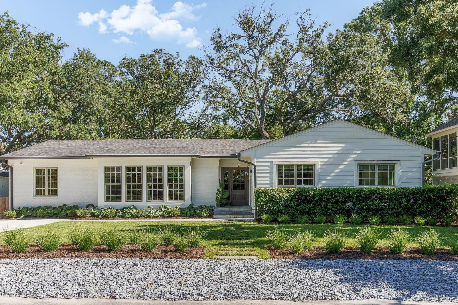White single-story house with broad windows, landscaped lawn, hedges, and mature trees.