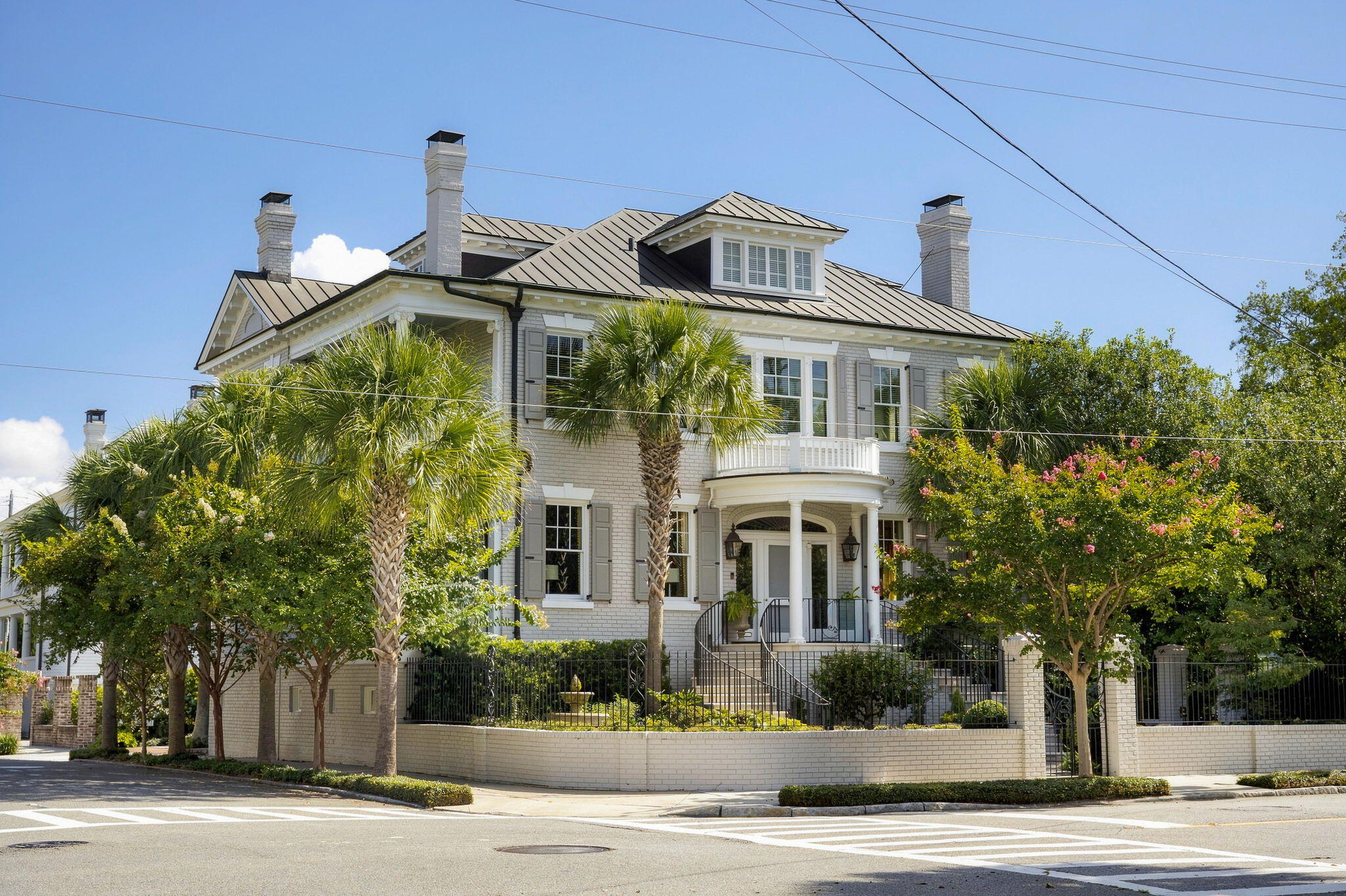 Two-story brick home with a metal roof, columned entrance, balcony, and palm-lined garden.