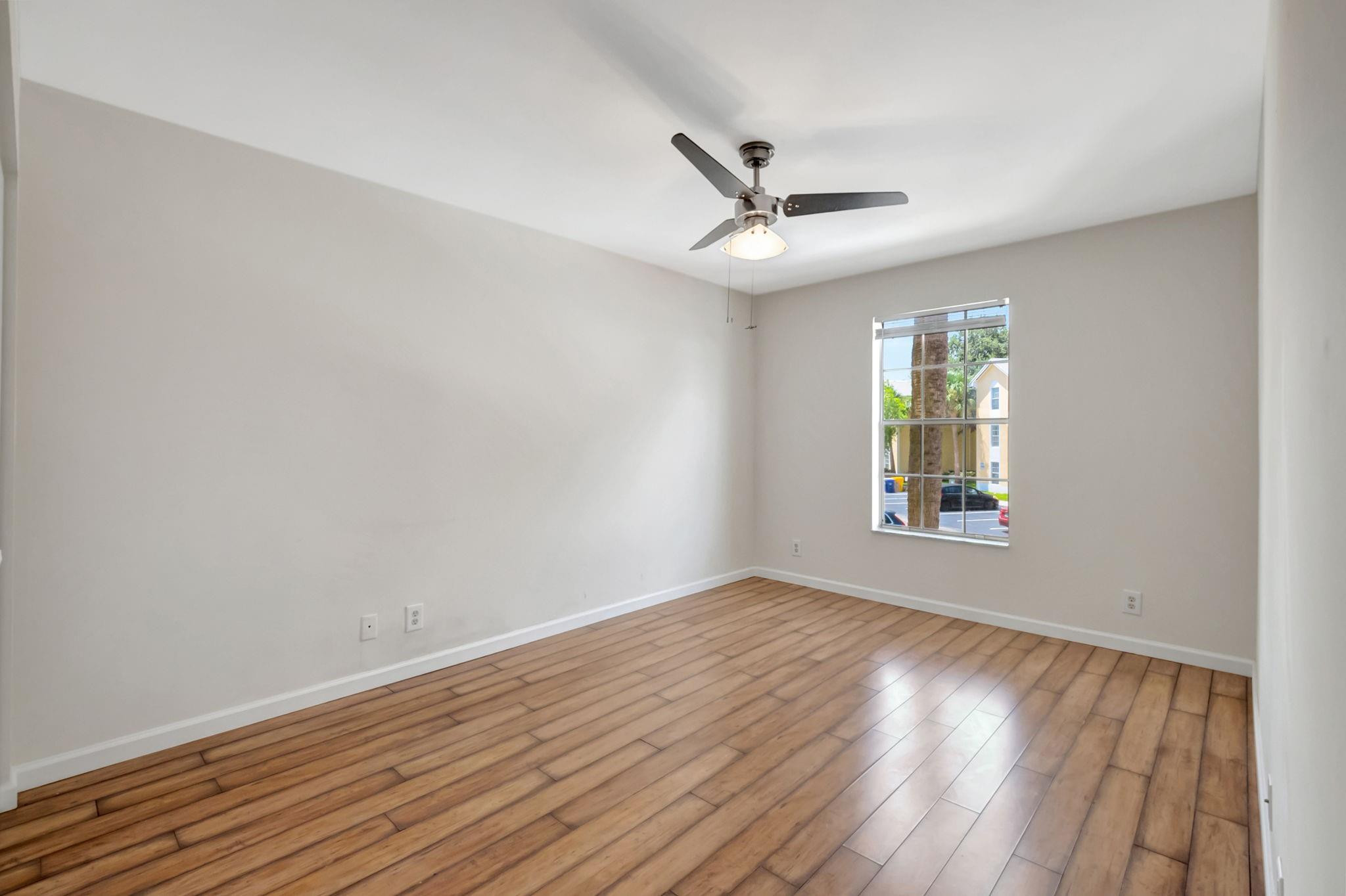 Empty bedroom with wood-look flooring, a ceiling fan, and a window overlooking neighboring buildings.