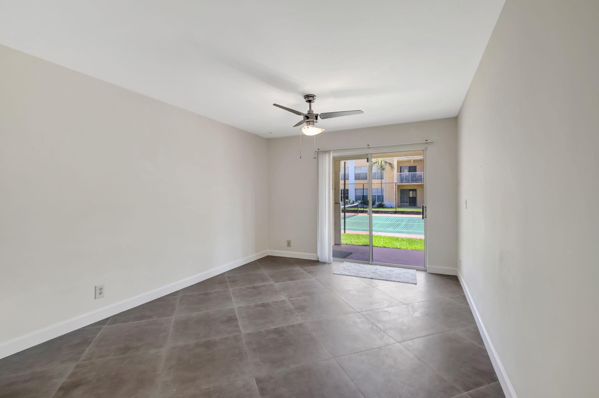 Vacant tiled room with a ceiling fan and sliding glass doors overlooking a tennis court.