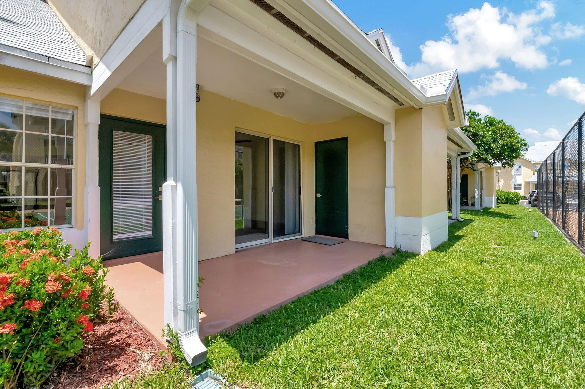 Home’s covered rear patio with sliding glass doors, green lawn, and flowering shrubs.