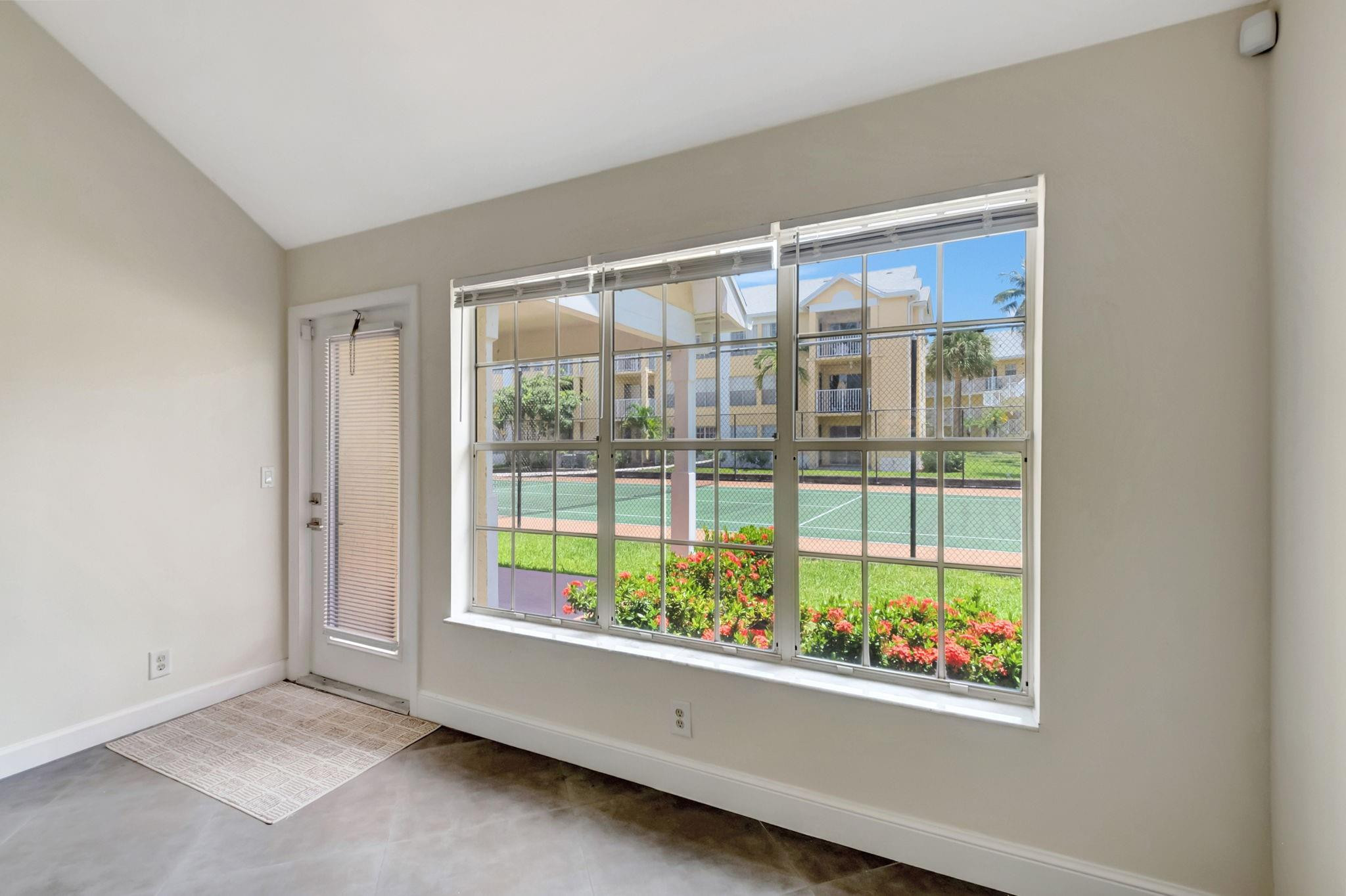 Empty room with large windows overlooking a tennis court and landscaped grounds.