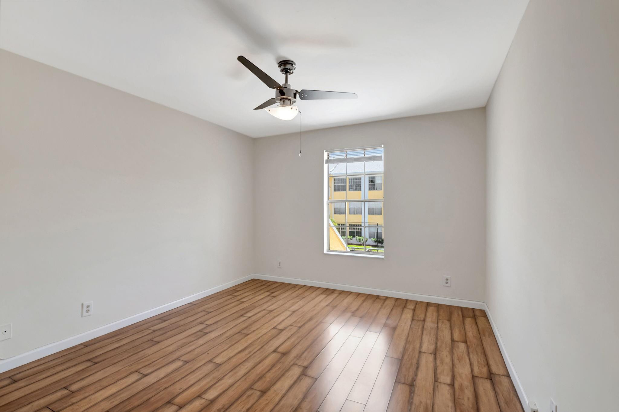Empty bedroom with wood-look flooring, ceiling fan, and a window overlooking a neighboring building.