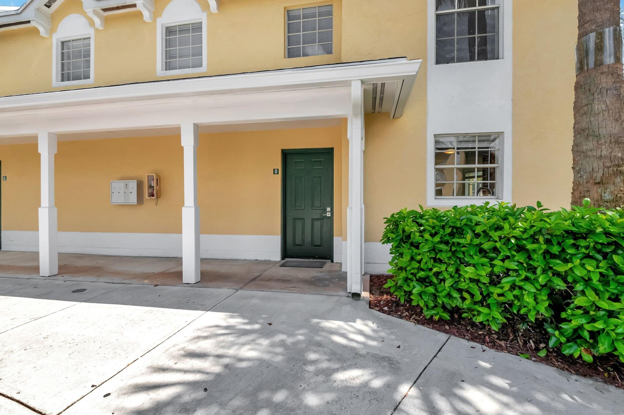 Unit D entrance beneath a covered walkway, with a green door, yellow stucco walls, and a trimmed hedge.