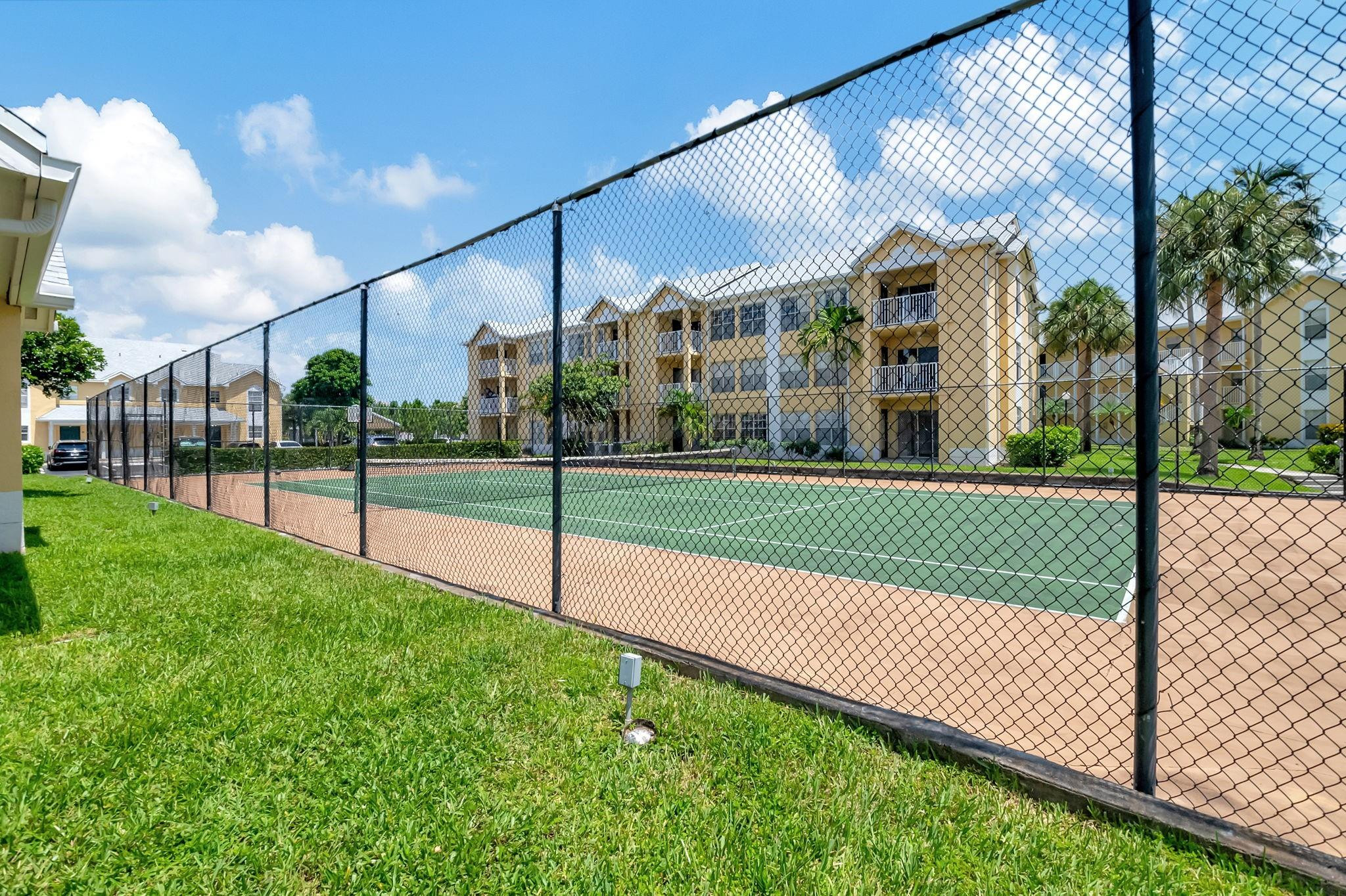 Fenced tennis court in front of beige condominium buildings and palm trees.