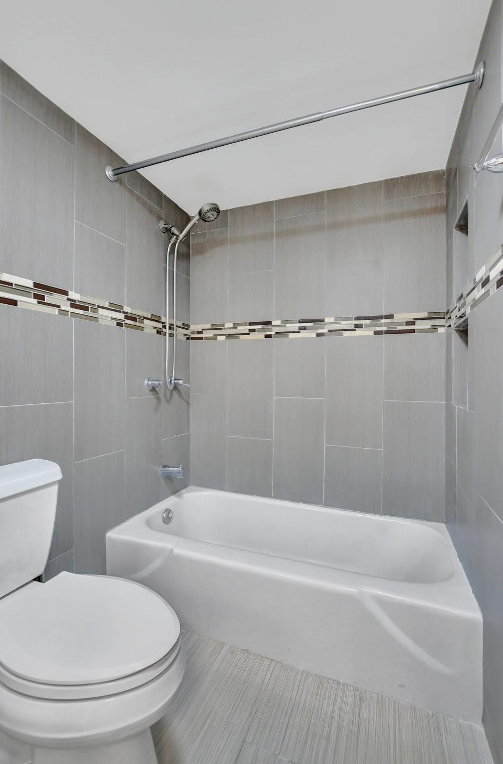 Bathroom with a white tub, handheld showerhead, gray tiles, and a narrow mosaic accent band.