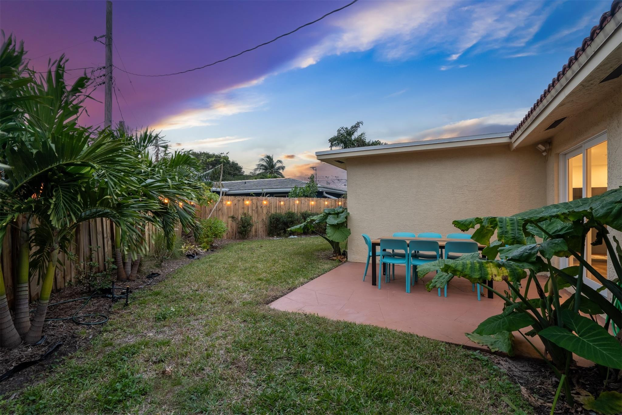 Backyard patio with turquoise dining chairs, tropical plants, and glowing fence lights at dusk.