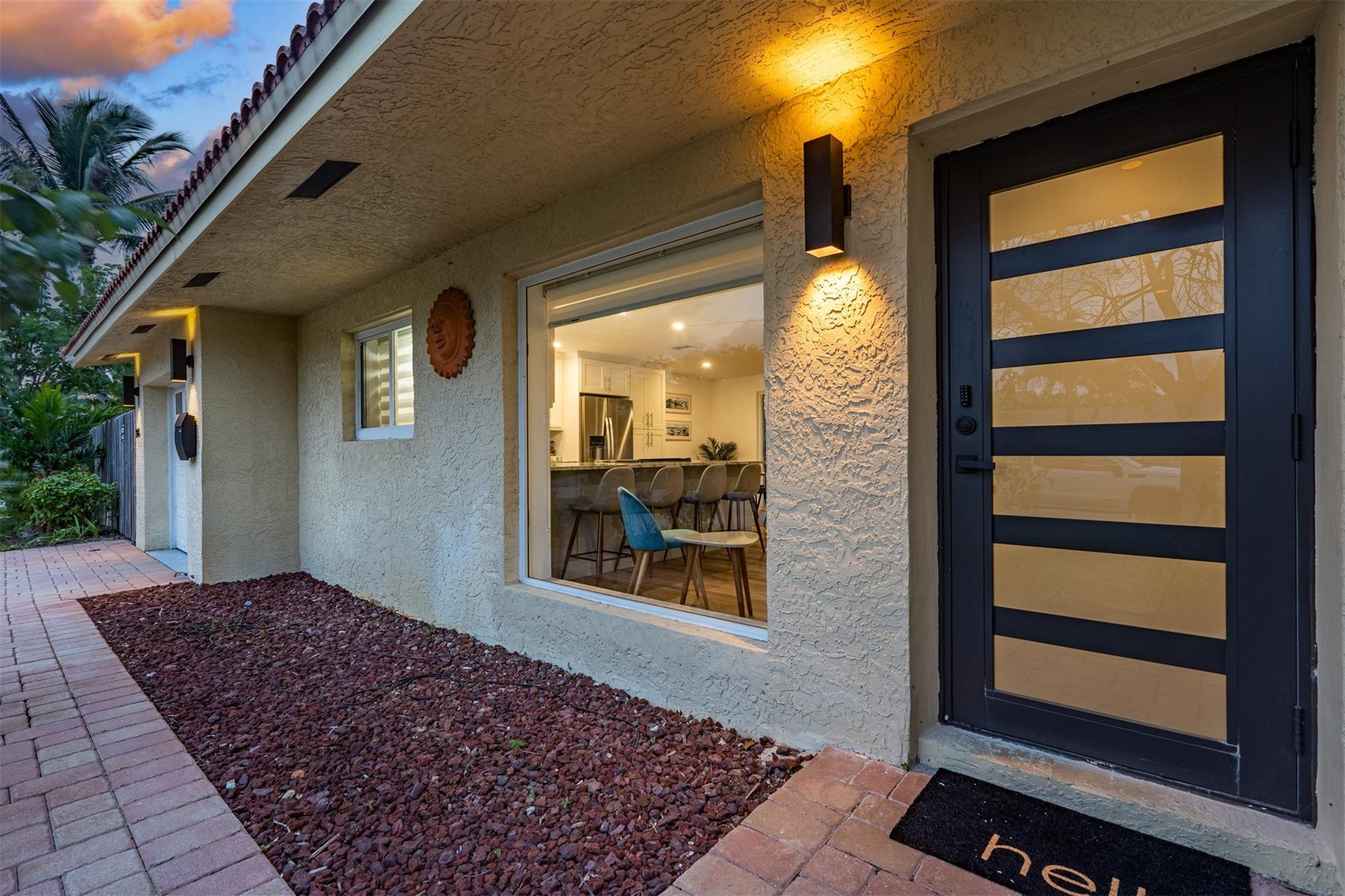 Home entrance with a black glass-panel door, illuminated stucco exterior, and paver walkway at dusk.