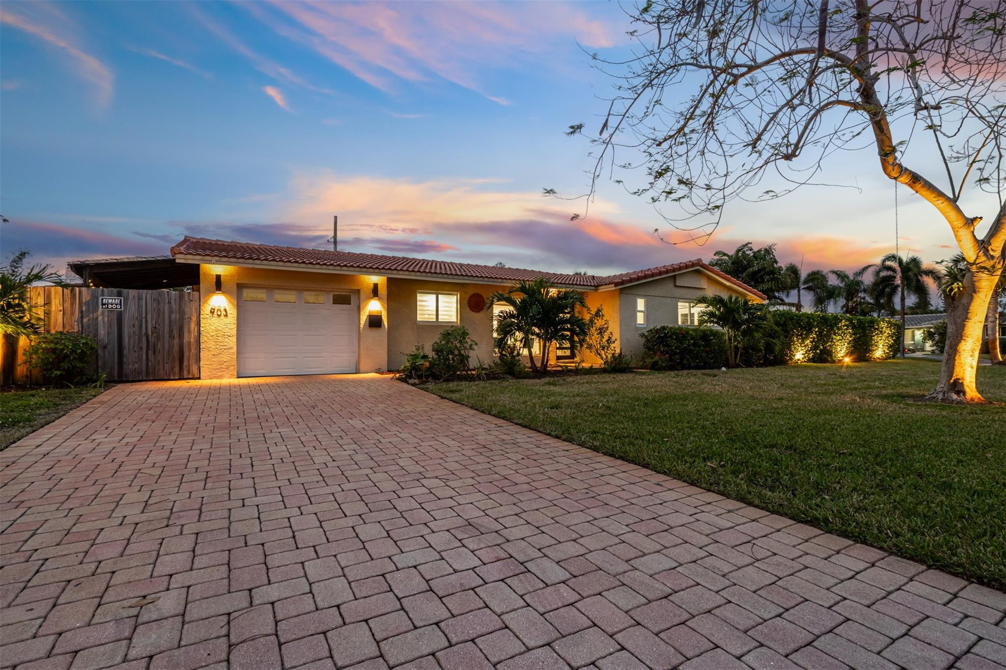 Single-story home with a red tile roof, illuminated facade, tropical landscaping, and broad paver driveway at dusk.