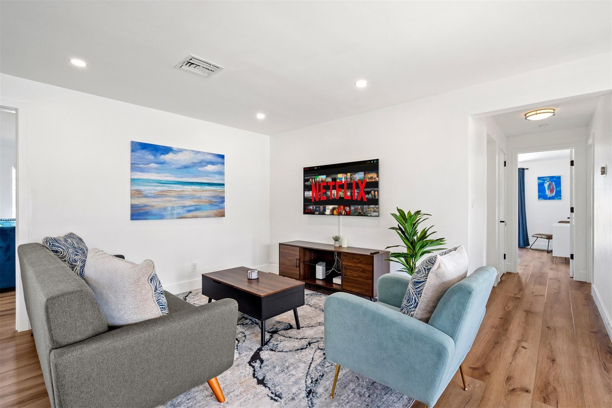 Furnished living room with gray and pale blue sofas, a wall-mounted television, and beach artwork.