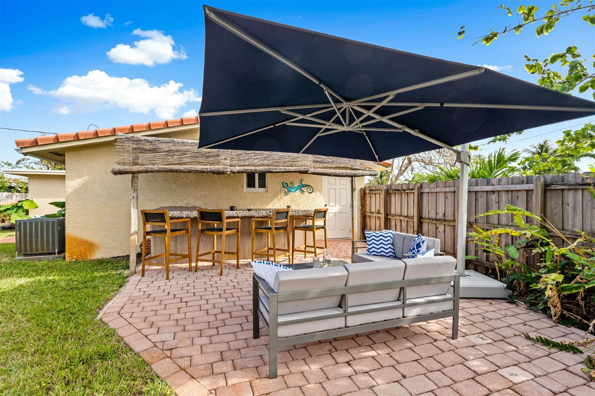 Backyard patio with brick pavers, cushioned seating, a large umbrella, and a bar counter.