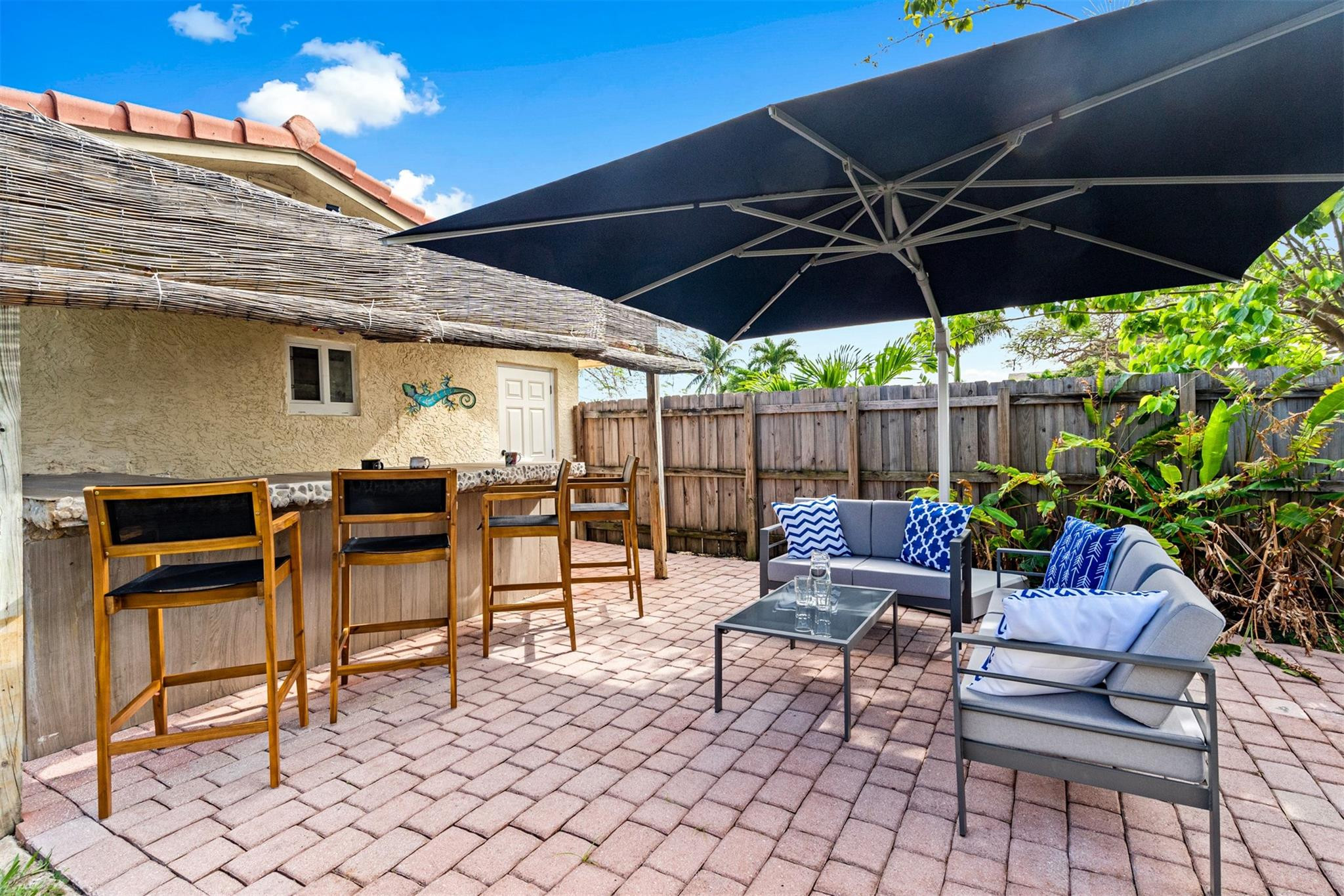 Backyard patio with brick pavers, oversized umbrella, lounge seating, and wooden bar chairs.